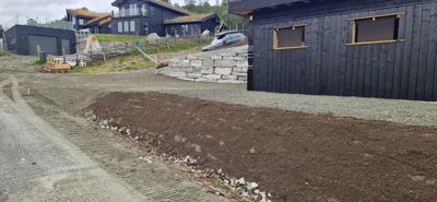 Fotografi av en byggeplass med et mørkt trehus i forgrunnen, en steinmur og andre hus på bakken.