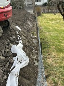 Fotografi av en utgravet terrengside med en hvit plastduk (muligens for å beskytte mot erosjon eller vann) og en steinmur langs en hekk.