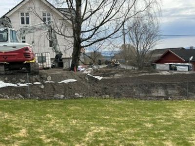 Fotografi av et byggefelt med en grøft, en grøftmaskin og en hvit bygning i bakgrunnen.