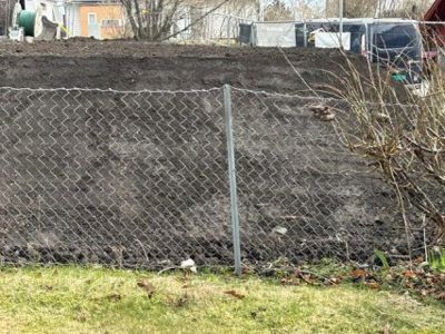 Fotografi av en grøft eller utgravet område bak en hekk, med en bil og en bygning i bakgrunnen.