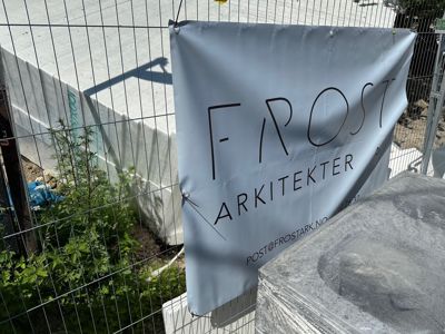 Fotografi av en grå banner med logoen 'FROST ARKITEKTER' hengt opp på en gjerde ved en byggeplass.