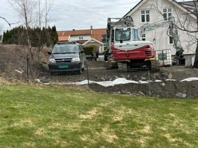 Fotografi av en byggeplass med en grøft, en bil og en graver som er i ferd med å grave ut en ny tomt foran en eksisterende hvit villa.